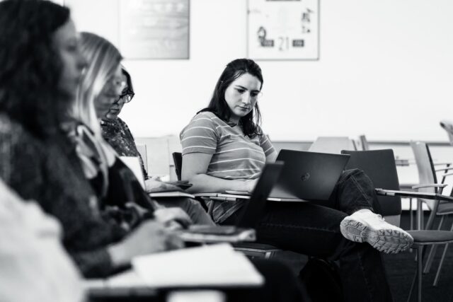 Kobieta pracująca na laptopie w sali, z przodu grupa osób