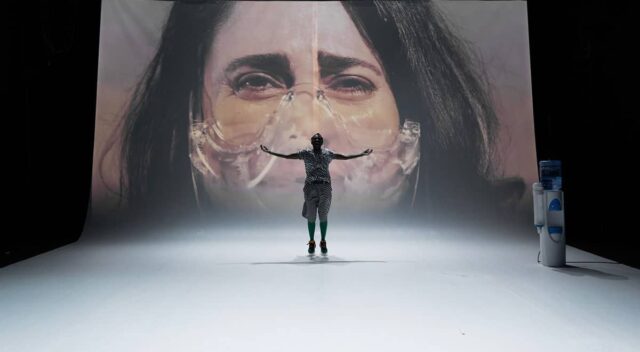 czarnoskóra kobieta na środku sceny ze zdjęciem innej kobiety na ekranie w tle