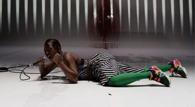 Czarnoskóra kobieta w stroju w czarno-białe paski, zielonych rajstopach leżąca na białej podłodze z mikrofonem w ręku