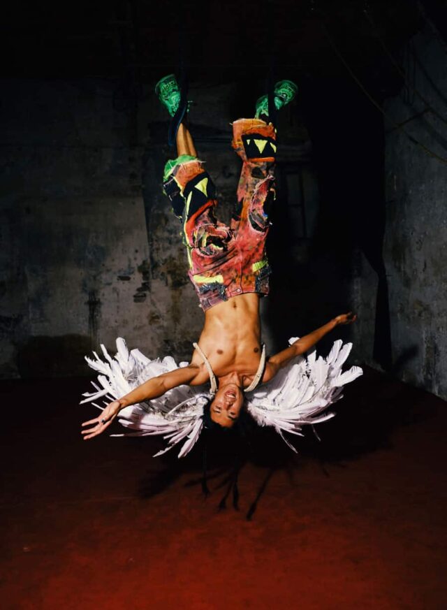 Męzczyzna w kolorwych spodniach z piórami wiszący głową w dół