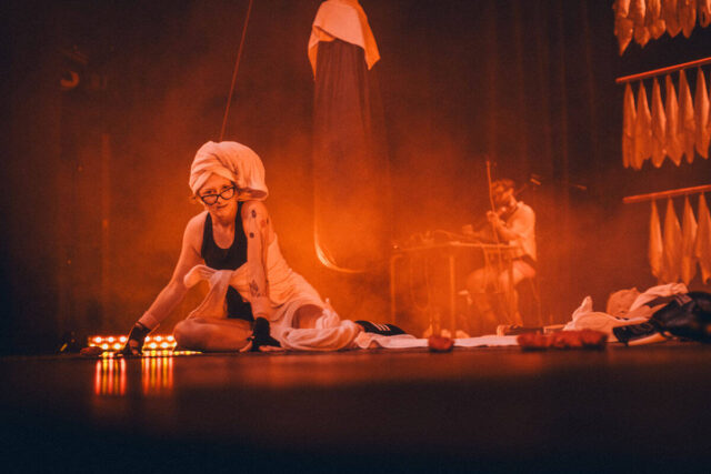 Kobieta w okularach z ręcznikiem na głowie patrzy w obiektyw aparatu. Czerwone światło na scenie.W tle wisi worek treningowy. Z tyłu siedzi osoba i gra na skrzypcach.