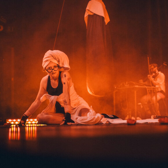 Kobieta w okularach z ręcznikiem na głowie patrzy w obiektyw aparatu. Czerwone światło na scenie.W tle wisi worek treningowy. Z tyłu siedzi osoba i gra na skrzypcach.