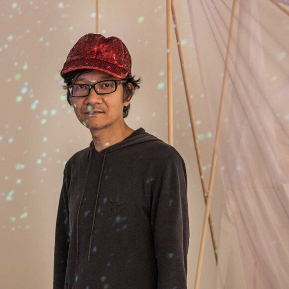 Artysta w czapce z daszkiem i okularach na tle wiszących tkanin