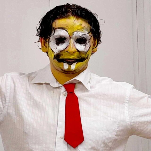Mlody mężczyzna w białej koszuli z czerwonym krawatem i pomalowana na żółto twarzą jak klaun