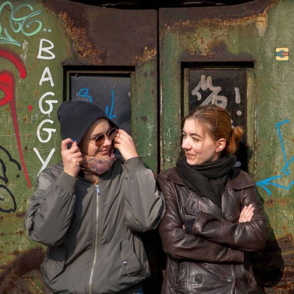 Dwie nastolatki siedzą na tle ściany z graffiti i patrzą na siebie