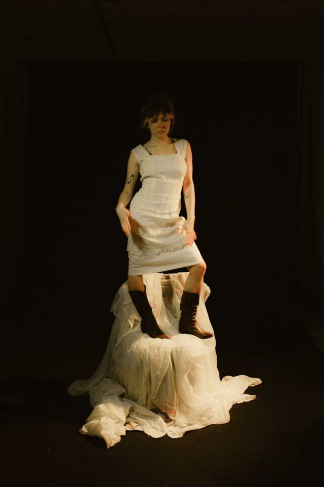 Młoda kobieta w białej sukni stojąca na podwyższeniu na czarnym tle