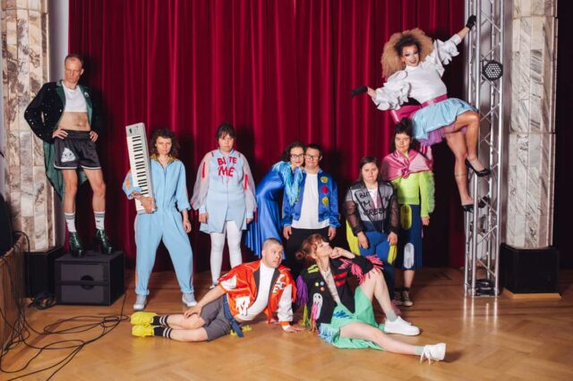 Aktorzy spektaklu w kolorowych strojach na tle czerwonej kotary