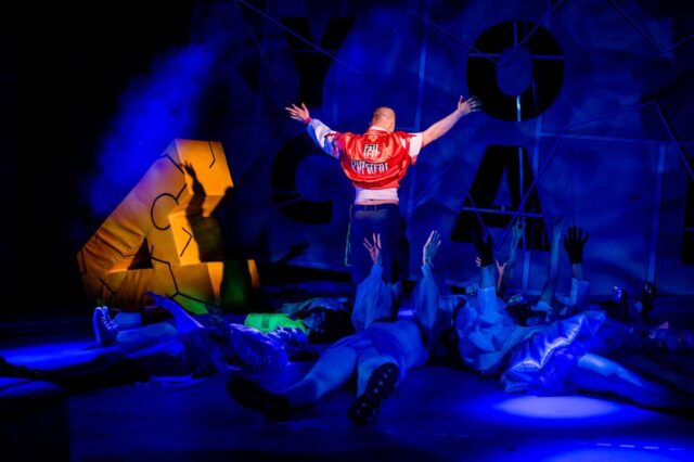 Mężczyzna w czerwonej kurtce stoi tyłem podnosi ręce do góry, na podłodze leżą inni ludzie słabo widoczni