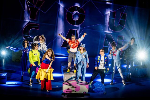 9 aktorów kolorowo ubranych na scenie, za nimi napis YOU CAN