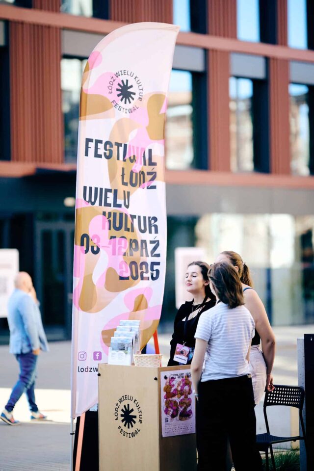 3 młode kobiety stoją przy ladzie z logo festiwalu, w tle flaga reklamowa i zabudowania