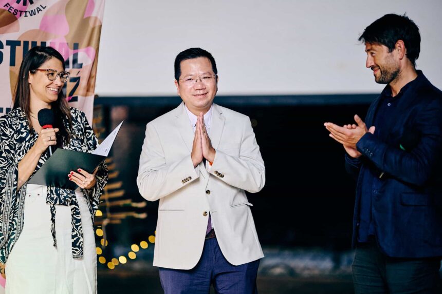 3 osoby: po lewej kobieta z mikrofonem, mężczyzna Wietnamczyk w centrum, po prawej mężczyzna bijący brawo