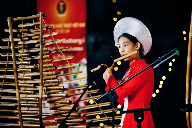 Kobieta w czerwonej sukni i białym nakryciu głowy gra na flecie, przed nią tradycyjny instrument