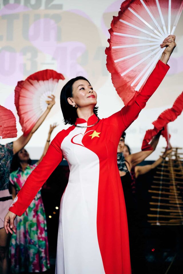 Wietnamka w biało-czerwonej sukni z wachlarzem, w tle inne osoby z wachlarzami