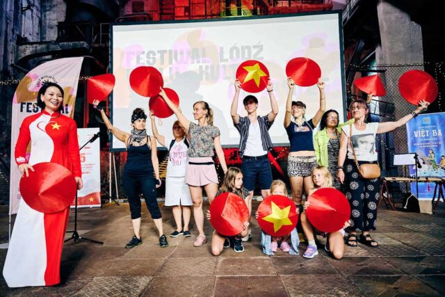 Grupa ludzi z czerwonymi tradycyjnymi nakryciami głowy w dłoniach, po prawej stronie Wietnamka w tradycyjnym stroju