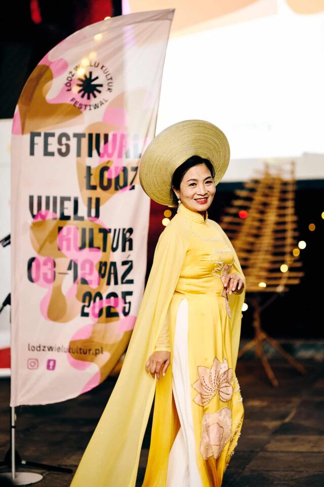 Wietnamka w tradycyjnej sukni i nakryciu głowy stoi przy fladze reklamującej Festiwal Łódź Wielu Kultur