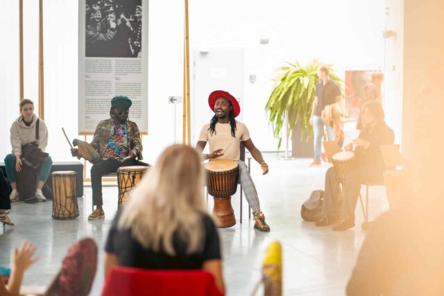 Czarny mężczyzna w czerwonym kapeluszu gra na bębnie, dookoła siedzą ludzie
