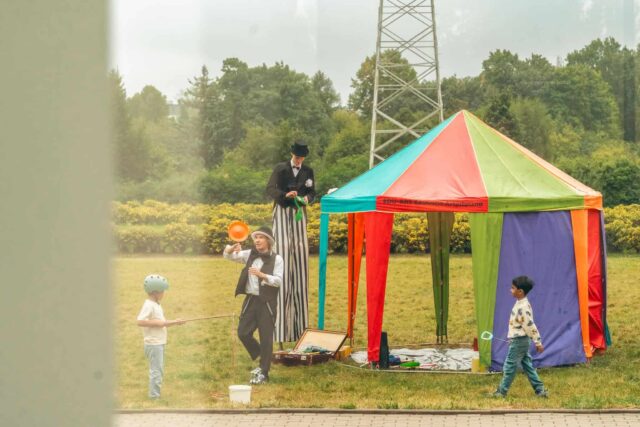 Mały namiot cyrkowy, mężczyzna z szczudłach i bawiące się dzieci