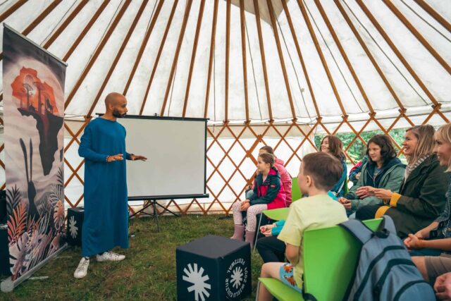 Mężczyzna w długiej szacie stoi przed grupą osób we wnętrzu namiotu jurty