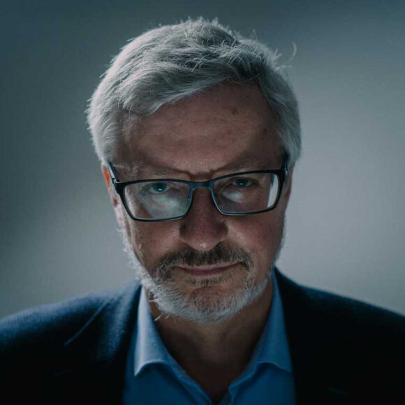 Siwy mężczyzna z brodą w okularach, w koszuli i marynarce patrzy w obiektyw