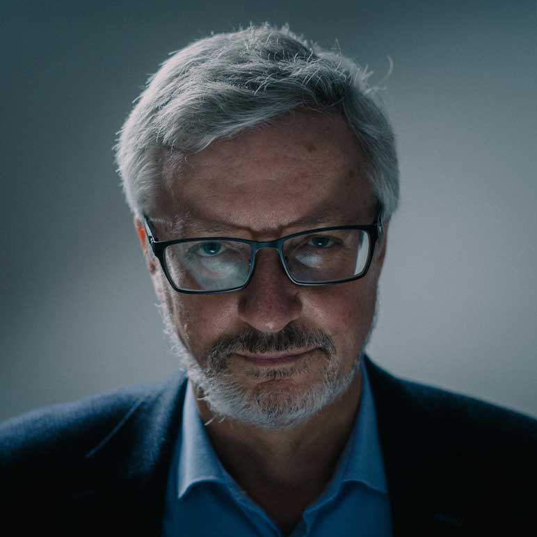 Siwy mężczyzna z brodą w okularach, w koszuli i marynarce patrzy w obiektyw