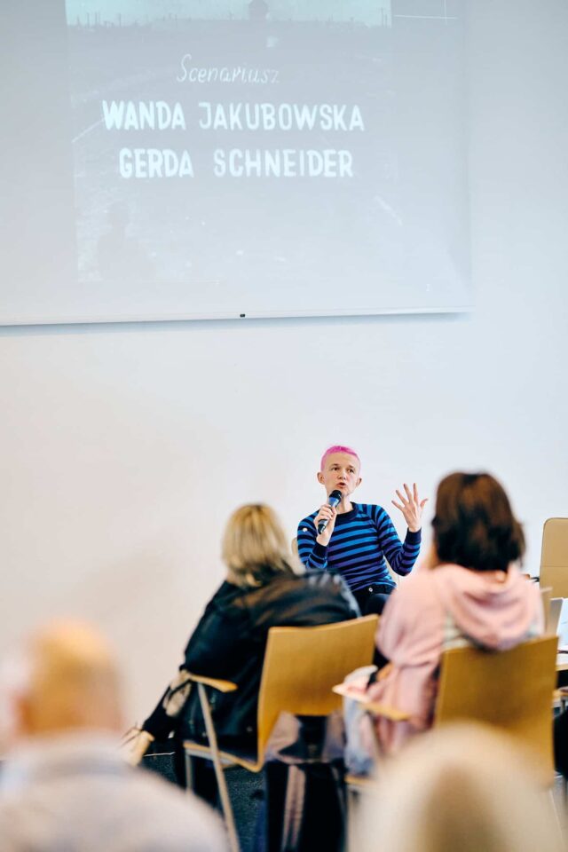 Kobieta z krótkimi różowymi włosami mówi do ludzi siedzących na krzesłach