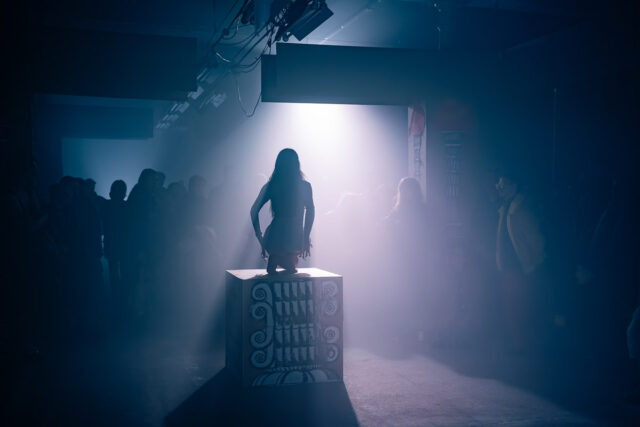 ciemne industrialne wnętrze, po środku sylwetka kobiety kucającej na podeście