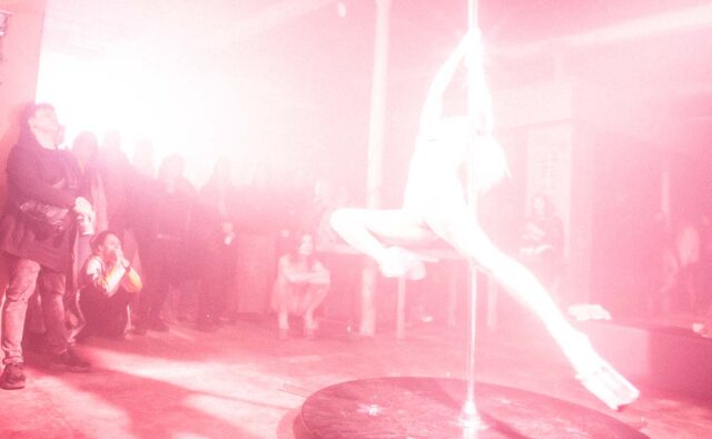 Różowe, prześwietlone zdjęcie z sylwetką kobiety tańczącą na rurze, publicznosc w tle