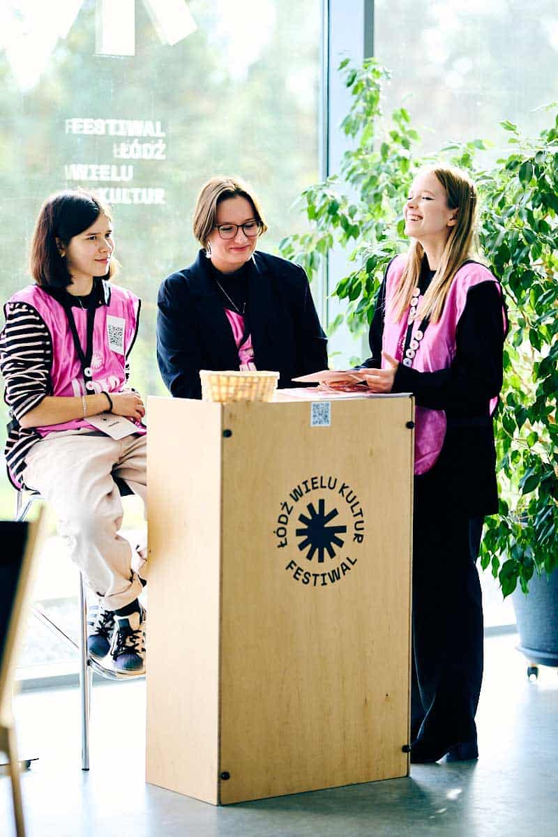 Trzy wolontariuszki w różowych kamizelkach przy ladzie z napisem Festiwal Łódź Wielu Kultur