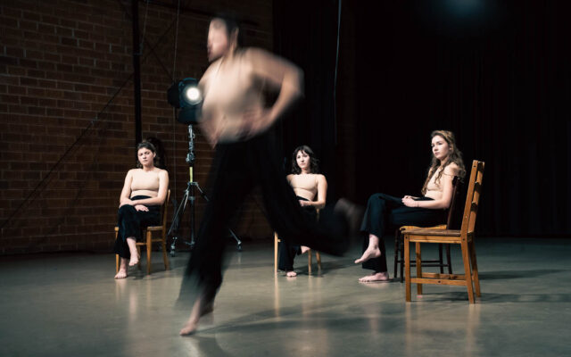trzy mlode kobiety siedzące na scenie na krzesłach, na pierwszym planie rozmazana postać czwartej