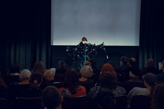 Kobieta przy mikrofonie owinięta czarną taśmą filmową, w tle ekran na pierwszym planie publiczność