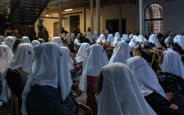 osoby - widzowie siedzą na krzesłach w białych nakryciach głowy z wyciętymi otworami na oczy