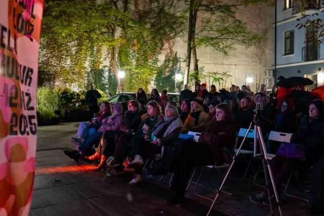 Publiczność siedząca na krzesłach na podwórku, na pierwszym planie flaga festiwalu