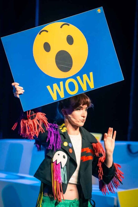 kobieta w kolorowym stroju podnosi planszę z emotikonką i napisem WOW